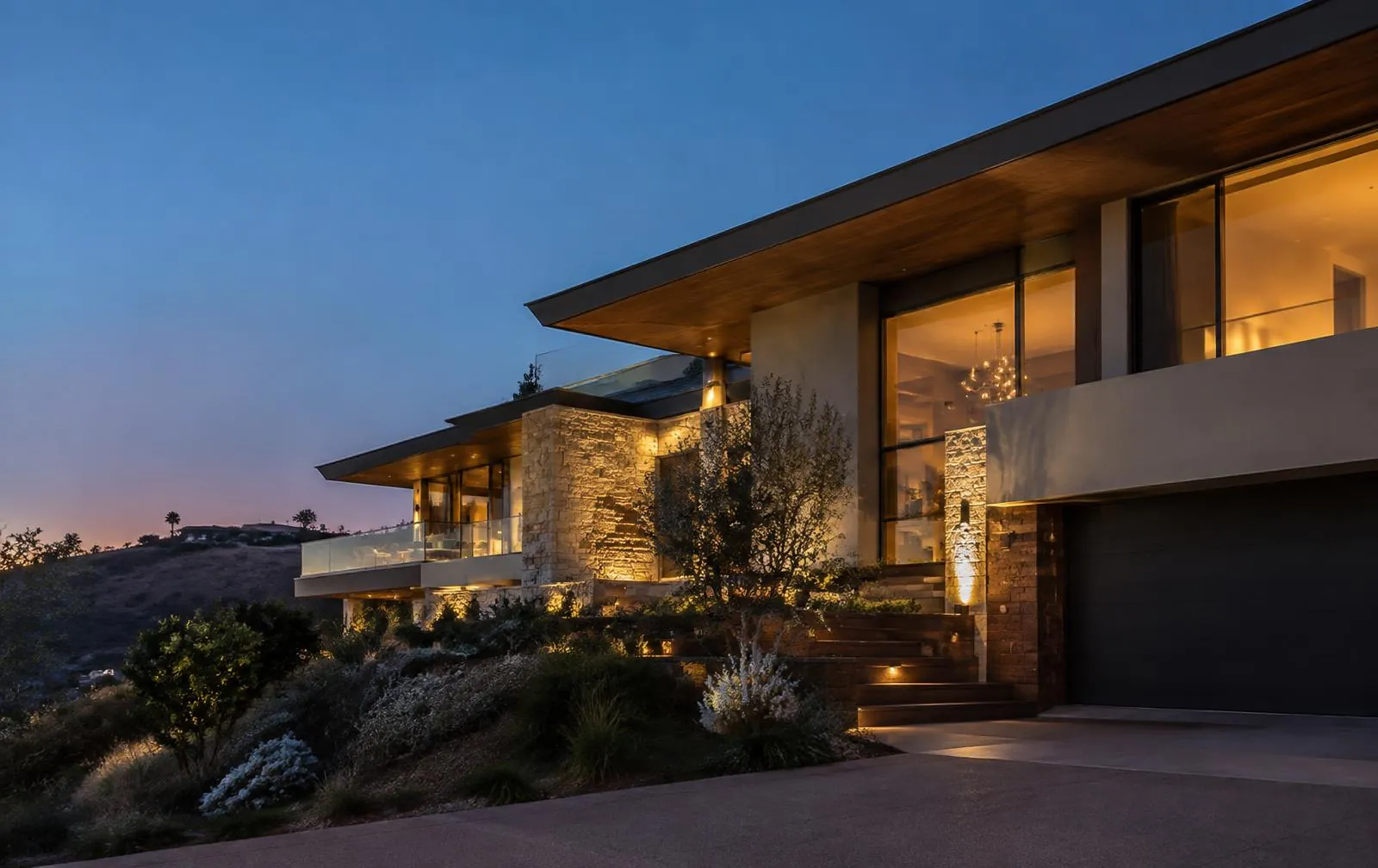 Contemporary custom hillside home at dusk with warm interior light, stone, and deep roof overhangs